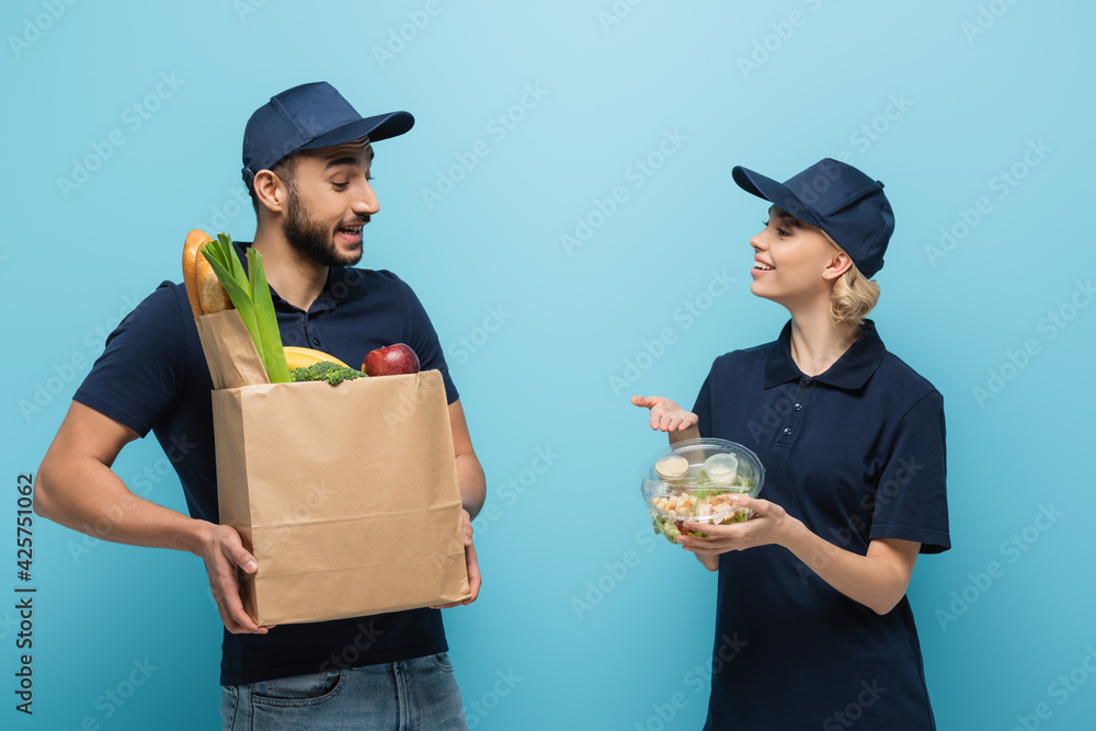 joyful courier pointing at lunch box near arabian colleague with food in paper bag on blue joyful courier pointing at lunch box near arabian colleague with food in paper bag on blue