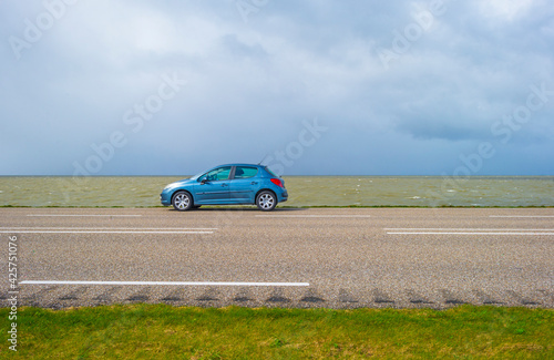 Car on a dike defying a stormy lake below a blue sky and white gray clouds in spring, Almere, Flevoland, The Netherlands, April 5, 2021
