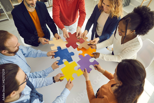 Diverse team connecting puzzle pieces as metaphor for finding solution to problem. High angle shot of happy business people putting together colorful jigsaw parts as symbol of teamwork and cooperation