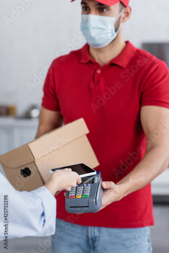 arabian delivery man in medical mask holding payment terminal near woman with smartphone, blurred background