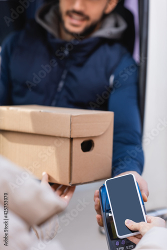 cropped view of woman holding parcel and paying with smartphone on terminal near postman on blurred background