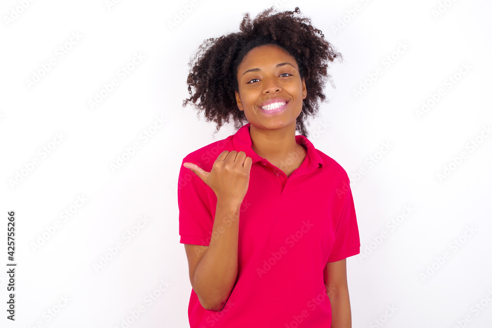Stupefied young beautiful African American woman wearing pink t-shirt with surprised expression, opens eyes and mouth widely, points aside with thumb, shows something strange. Advertisement concept.