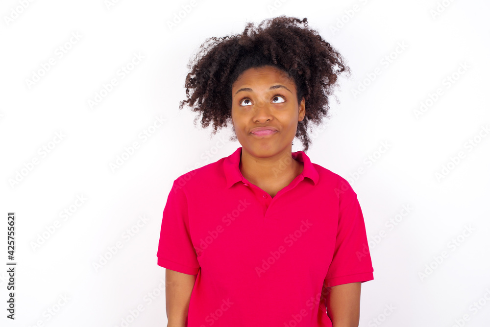 young beautiful African American woman wearing pink t-shirt against white wall crosses eyes, puts lips, makes grimace with awkward expression has fun alone, plays fool. young beautiful African American woman wearing pink t-shirt against white wall crosses eyes, puts lips, makes grimace with awkward expression has fun alone, plays fool.