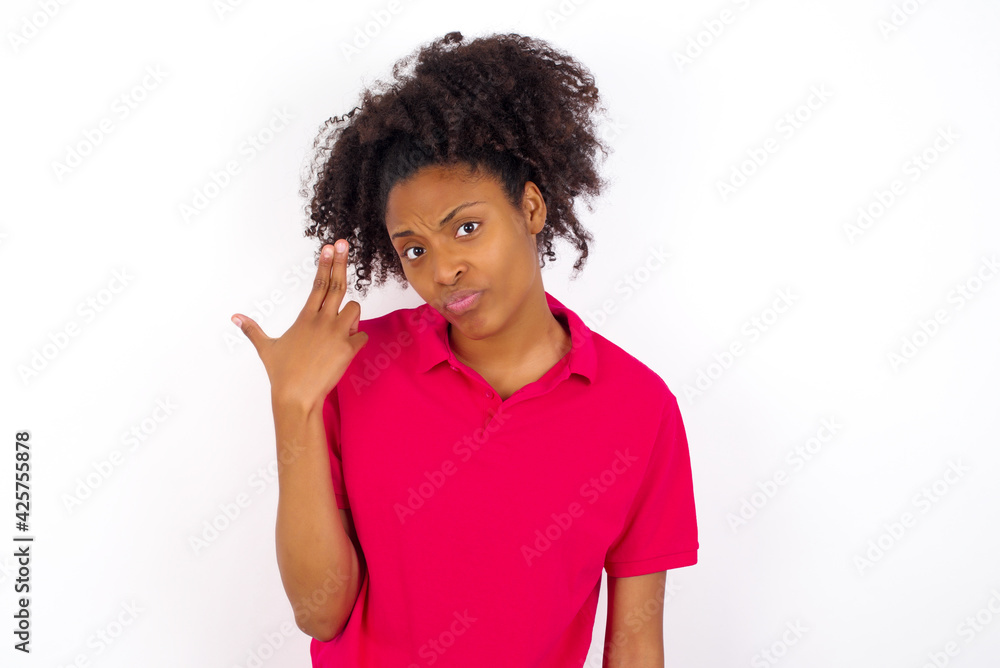 Unhappy young beautiful African American woman wearing pink t-shirt against white wall imitates gun shoot makes suicide gesture keeps two fingers on temples. Unhappy young beautiful African American woman wearing pink t-shirt against white wall imitates gun shoot makes suicide gesture keeps two fingers on temples.