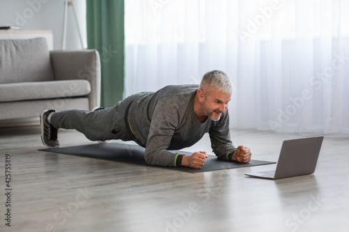 Athletic mature bearded man doing exercises at home, using laptop