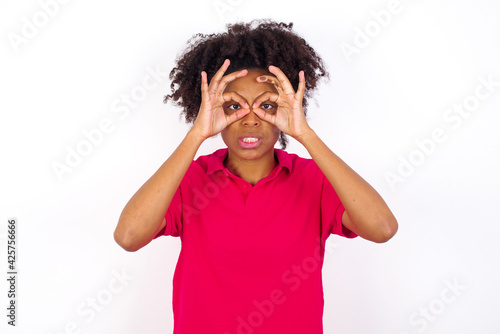 Playful excited young beautiful African American woman wearing pink t-shirt against white wall showing Ok sign with both hands on eyes  pretending to wear spectacles.
