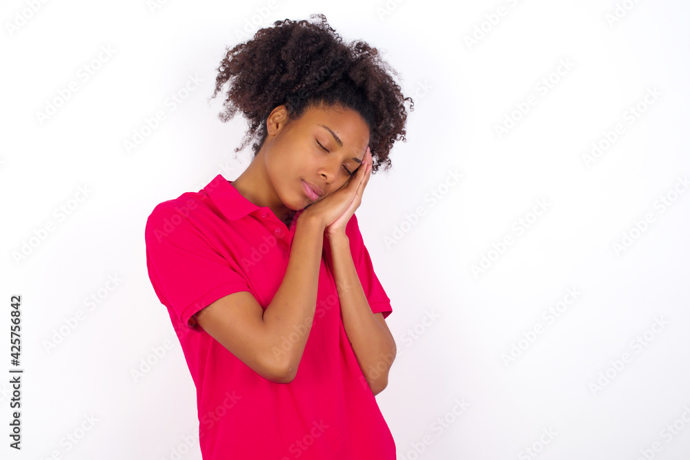 Relax and sleep time. Tired young beautiful African American woman wearing pink t-shirt against white wall with closed eyes leaning on palms making sleeping gesture. Relax and sleep time. Tired young beautiful African American woman wearing pink t-shirt against white wall with closed eyes leaning on palms making sleeping gesture.