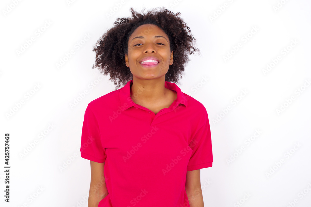 young beautiful African American woman wearing pink t-shirt against white wall with happy and funny face smiling and showing tongue.