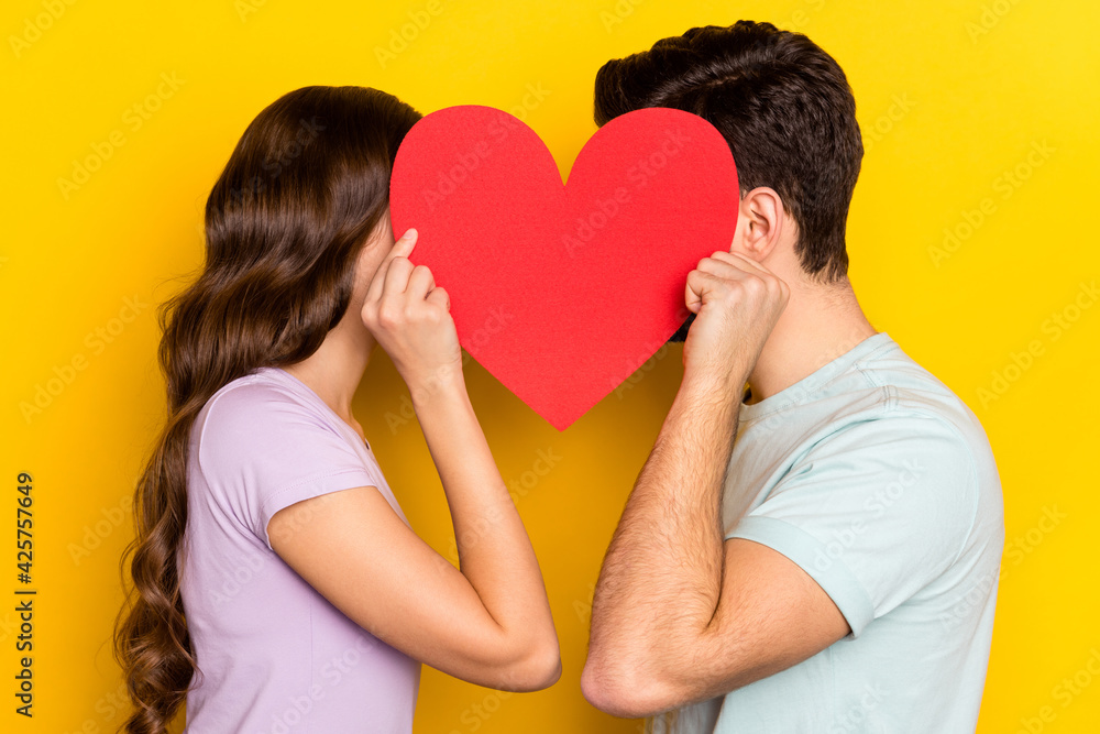 Profile side view portrait of couple holding big heart hiding face romance kissing isolated over bright yellow color background Profile side view portrait of couple holding big heart hiding face romance kissing isolated over bright yellow color background