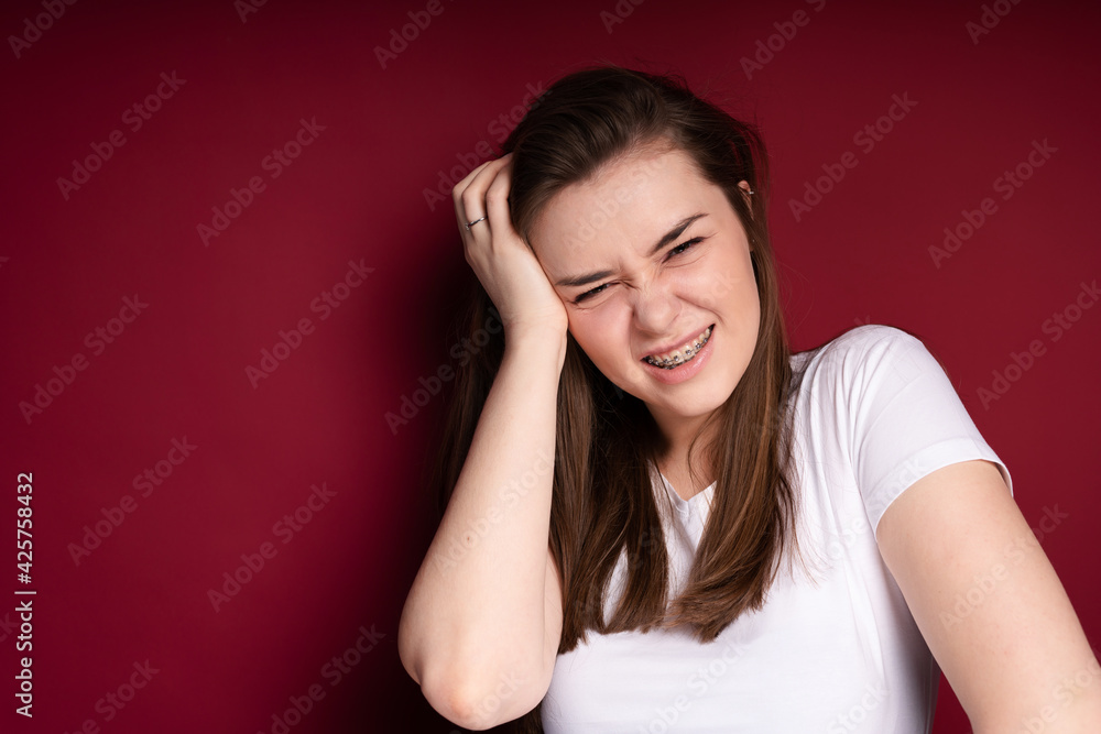 A young brunette tilts her head to the side, frowns in pain and touches her hand to her head. Pain after installing braces A young brunette tilts her head to the side, frowns in pain and touches her hand to her head. Pain after installing braces