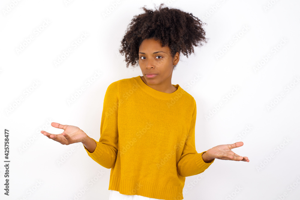 young beautiful African American woman wearing yellow sweater against white wall looks uncertain shrugs shoulders. young beautiful African American woman wearing yellow sweater against white wall looks uncertain shrugs shoulders.