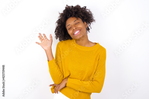 Overjoyed successful young beautiful African American woman wearing yellow sweater against white wall raises palm and closes eyes in joy being entertained by friends