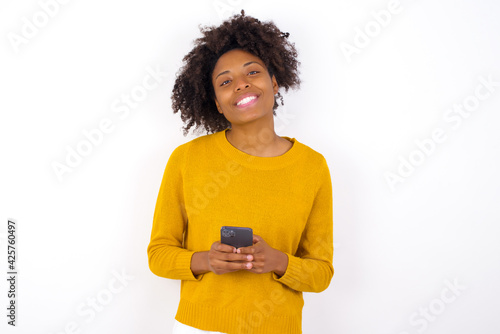 young beautiful African American woman wearing yellow sweater against white wall Mock up copy space. Using mobile phone, typing sms message