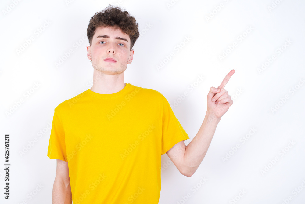 young caucasian handsome man with curly hair wearing yellow T-shirt against white studio background looking at camera indicating finger empty space sales young caucasian handsome man with curly hair wearing yellow T-shirt against white studio background looking at camera indicating finger empty space sales