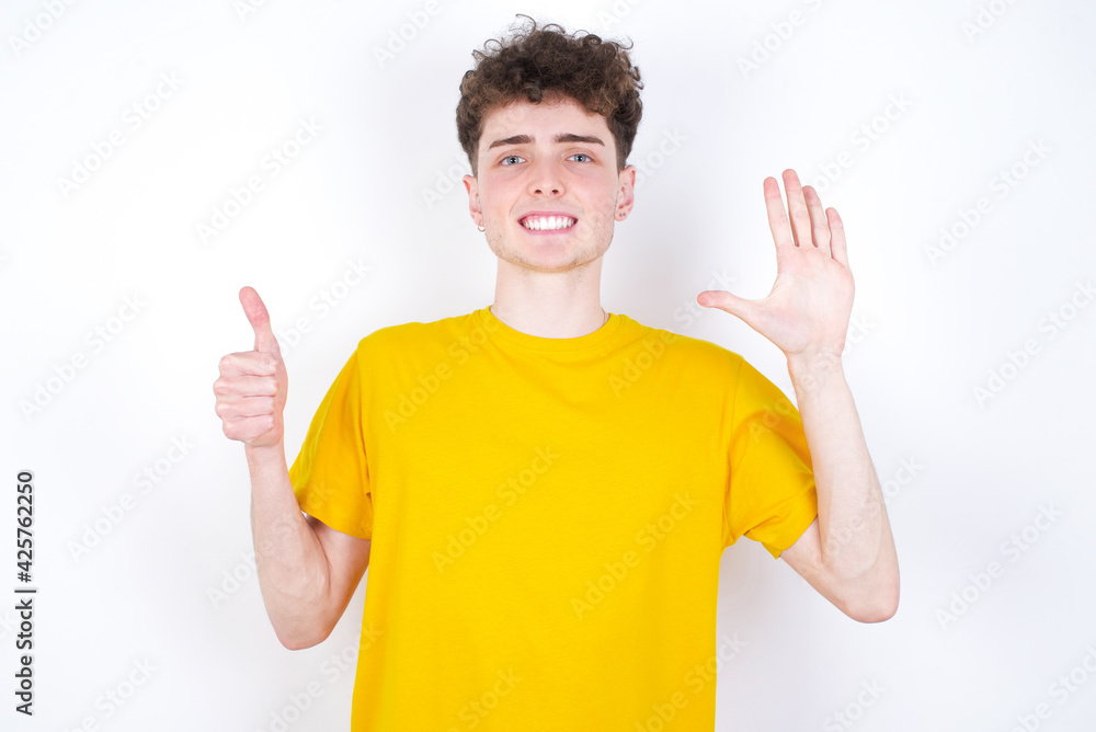 young caucasian handsome man with curly hair wearing yellow T-shirt against white studio background showing and pointing up with fingers number six while smiling confident and happy. young caucasian handsome man with curly hair wearing yellow T-shirt against white studio background showing and pointing up with fingers number six while smiling confident and happy.