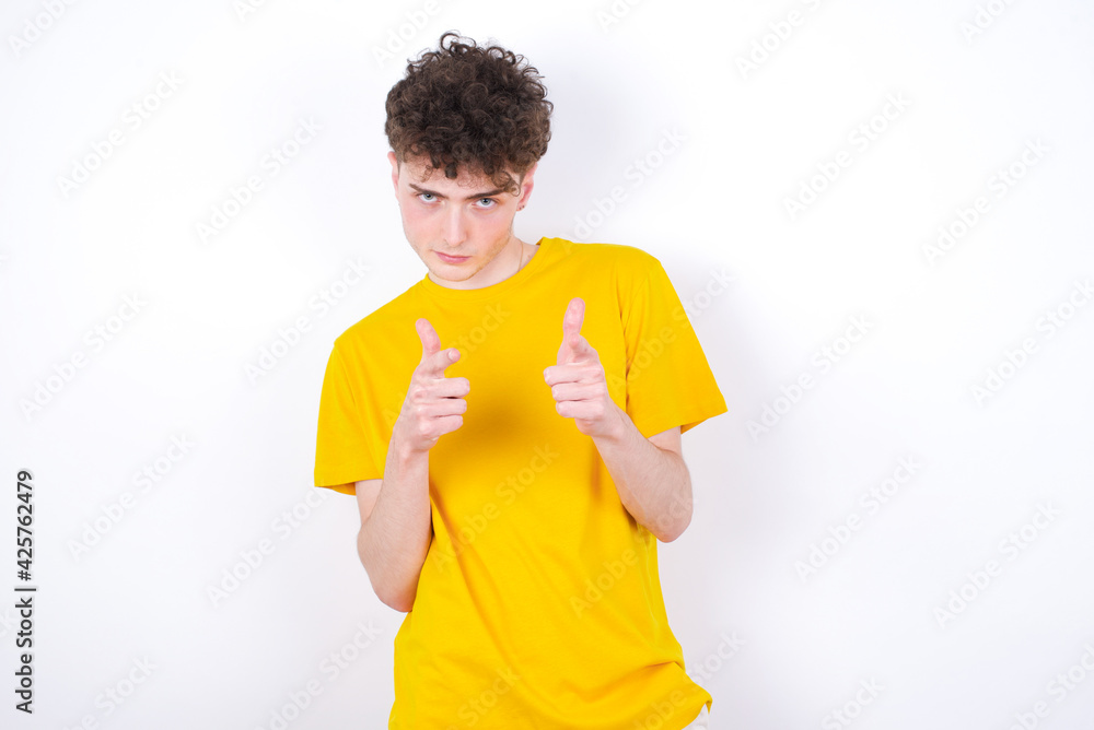 Joyful young caucasian handsome man with curly hair wearing yellow T-shirt against white studio background wink and points index fingers at camera, chooses someone, makes finger gun pistol. Joyful young caucasian handsome man with curly hair wearing yellow T-shirt against white studio background wink and points index fingers at camera, chooses someone, makes finger gun pistol.