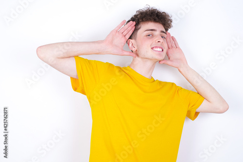 young caucasian handsome man with curly hair wearing yellow T-shirt against white studio background Trying to hear both hands on ear gesture, curious for gossip. Hearing problem, deaf