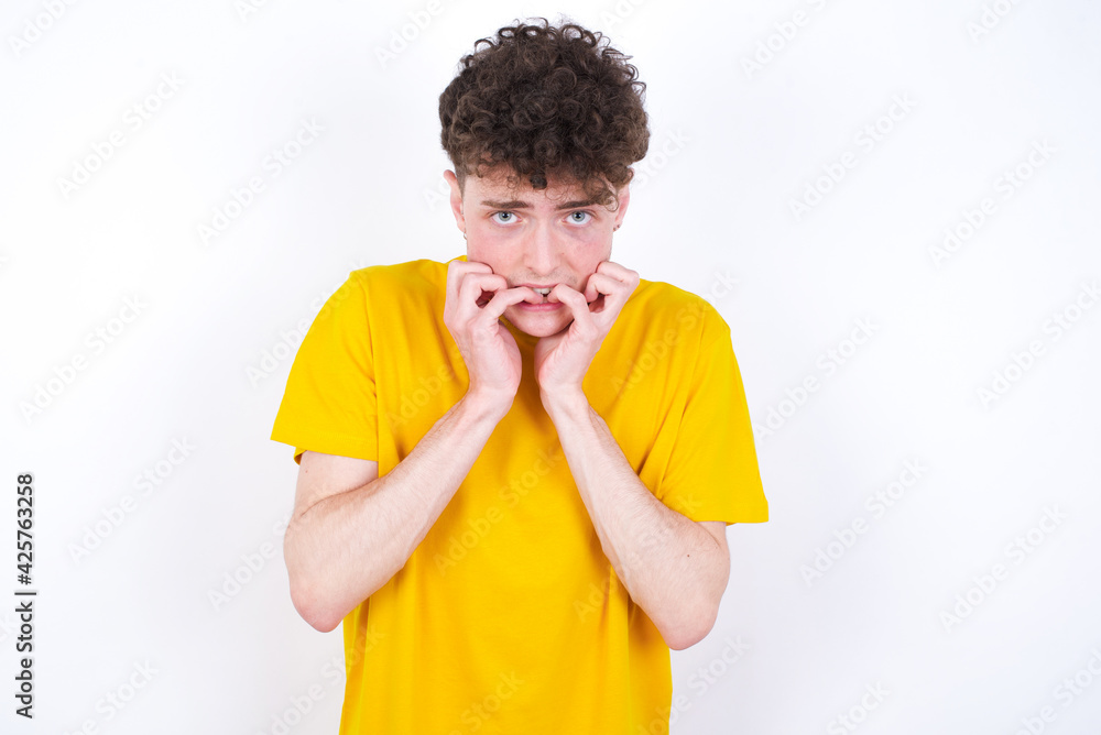 Fearful young caucasian handsome man with curly hair wearing yellow T-shirt against white studio wall keeps hands near mouth, feels frightened and scared, has a phobia, Shock and frighted concept. Fearful young caucasian handsome man with curly hair wearing yellow T-shirt against white studio wall keeps hands near mouth, feels frightened and scared, has a phobia, Shock and frighted concept.
