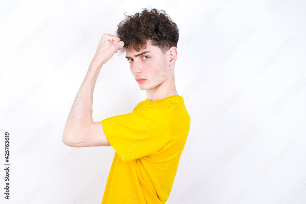 Portrait of powerful cheerful young caucasian handsome man with curly hair wearing yellow T-shirt against white studio background showing muscles. Portrait of powerful cheerful young caucasian handsome man with curly hair wearing yellow T-shirt against white studio background showing muscles.