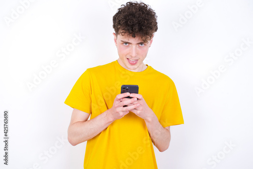 young caucasian handsome man with curly hair wearing yellow T-shirt enjoys distant communication, uses mobile phone, surfs fast unlimited internet, has pleasant smile, makes shopping online,