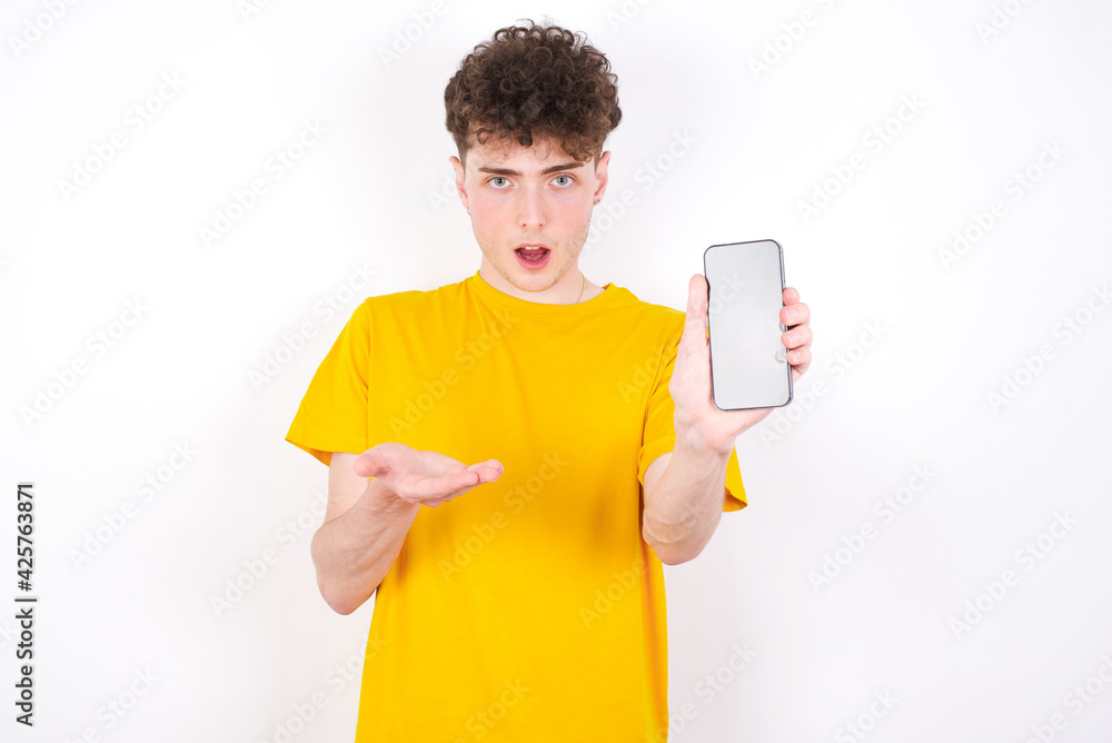 young caucasian handsome man with curly hair wearing yellow T-shirt against white studio background with a mobile. presenting smartphone. Advertisement concept. young caucasian handsome man with curly hair wearing yellow T-shirt against white studio background with a mobile. presenting smartphone. Advertisement concept.