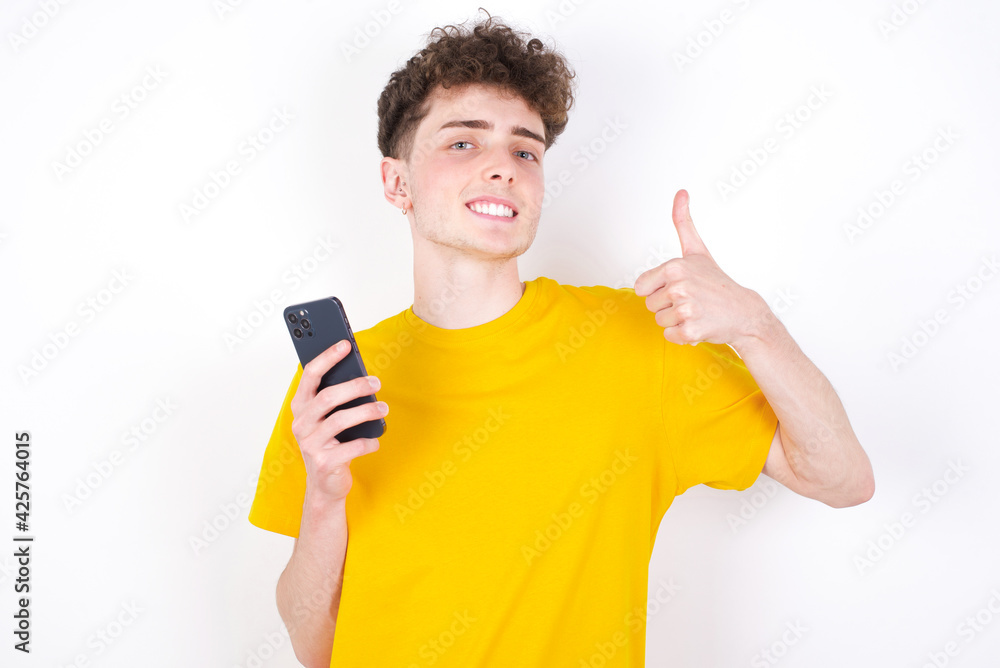 Portrait of young caucasian handsome man with curly hair wearing yellow T-shirt using and texting with smartphone happy with big smile doing ok sign, thumb up with fingers, excellent sign Portrait of young caucasian handsome man with curly hair wearing yellow T-shirt using and texting with smartphone happy with big smile doing ok sign, thumb up with fingers, excellent sign