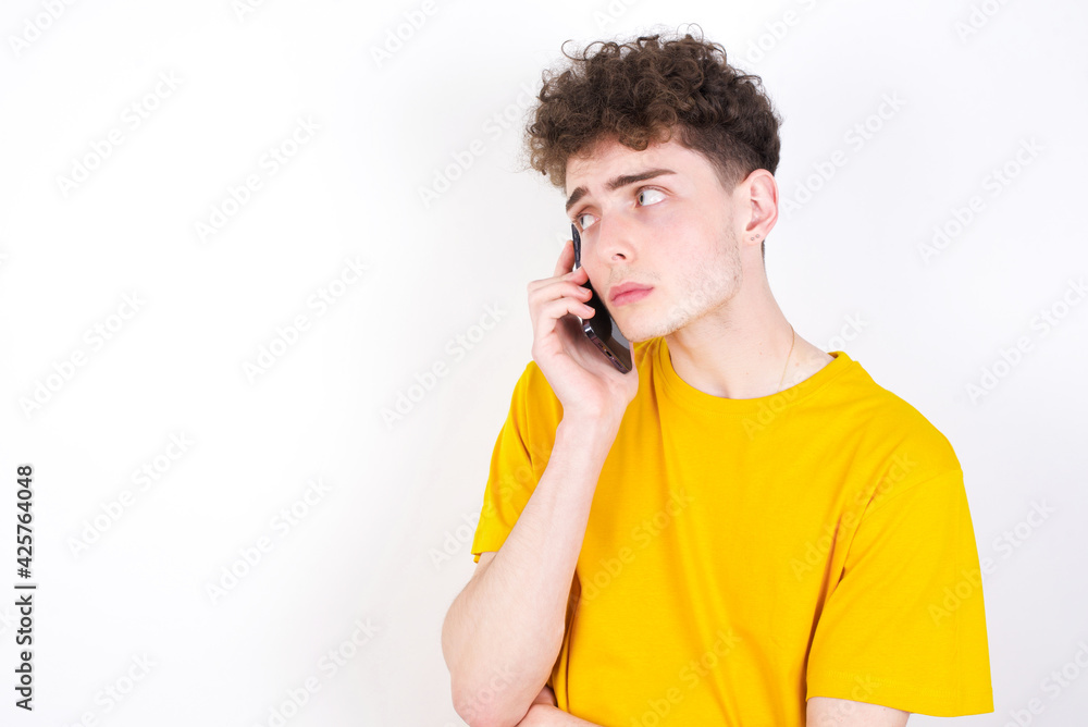 Sad young caucasian handsome man with curly hair wearing yellow T-shirt against white studio background talking on smartphone. Communication concept. Sad young caucasian handsome man with curly hair wearing yellow T-shirt against white studio background talking on smartphone. Communication concept.