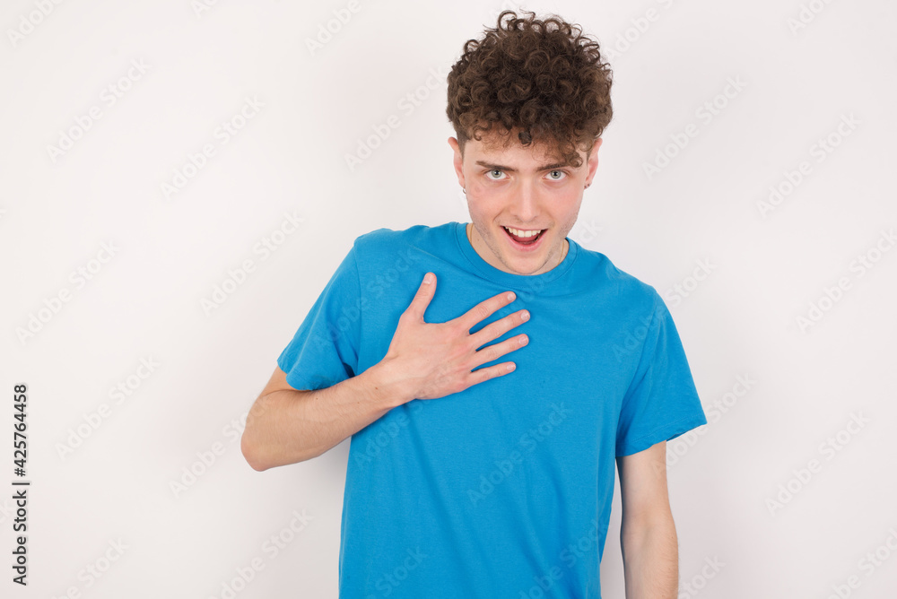 young caucasian handsome man with curly hair wearing blue T-shirt against white studio background smiles toothily cannot believe eyes expresses good emotions and surprisement young caucasian handsome man with curly hair wearing blue T-shirt against white studio background smiles toothily cannot believe eyes expresses good emotions and surprisement