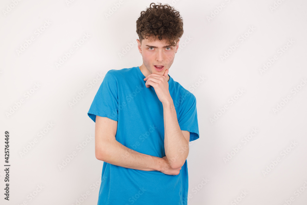 young caucasian handsome man with curly hair wearing blue T-shirt against white studio background laughs happily keeps hand on chin expresses positive emotions smiles broadly has carefree expression young caucasian handsome man with curly hair wearing blue T-shirt against white studio background laughs happily keeps hand on chin expresses positive emotions smiles broadly has carefree expression