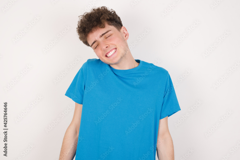 Positive young caucasian handsome man with curly hair wearing blue T-shirt against white studio background with overjoyed expression closes eyes and laughs shows white perfect teeth Positive young caucasian handsome man with curly hair wearing blue T-shirt against white studio background with overjoyed expression closes eyes and laughs shows white perfect teeth