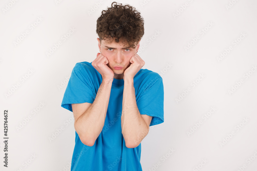 young caucasian handsome man with curly hair wearing blue T-shirt against white studio background with surprised expression keeps hands under chin keeps lips folded makes funny grimace young caucasian handsome man with curly hair wearing blue T-shirt against white studio background with surprised expression keeps hands under chin keeps lips folded makes funny grimace
