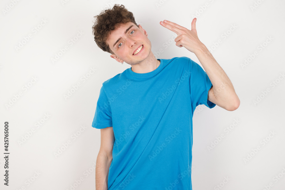 Fototapeta premium young caucasian handsome man with curly hair wearing blue T-shirt against white studio wall foolishes around shoots in temple with fingers makes suicide gesture. Funny model makes finger gun pistol