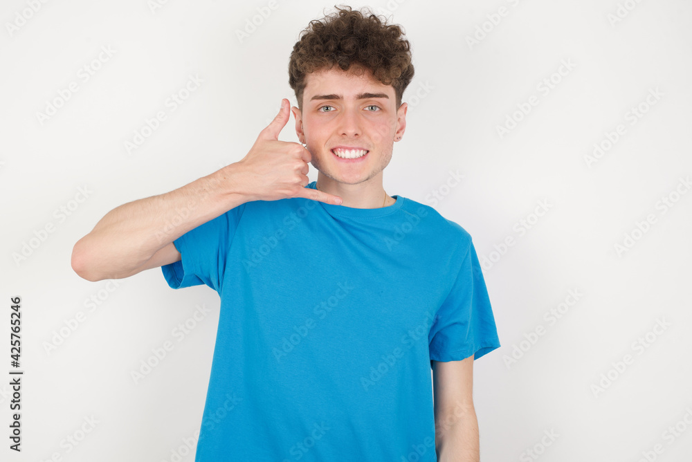 young caucasian handsome man with curly hair wearing blue T-shirt against white studio background makes phone gesture, says call me back again, has glad expression.