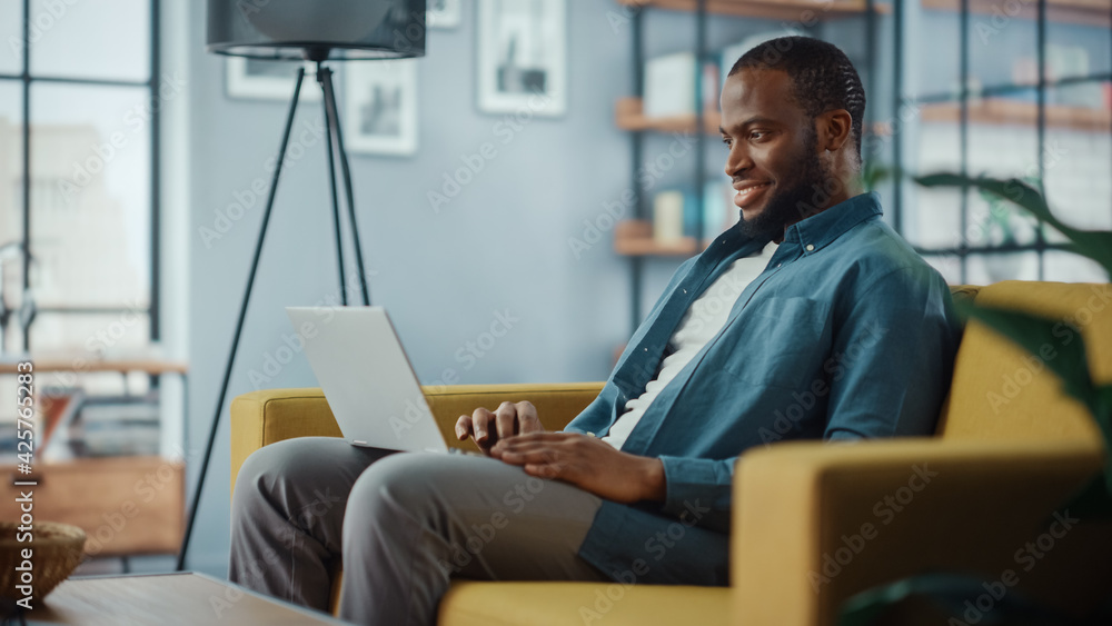 Handsome Black African American Man Working on Laptop Computer while Sitting on a Sofa in Cozy Living Room. Freelancer Working From Home. Browsing Internet, Using Social Networks, Having Fun in Flat.
