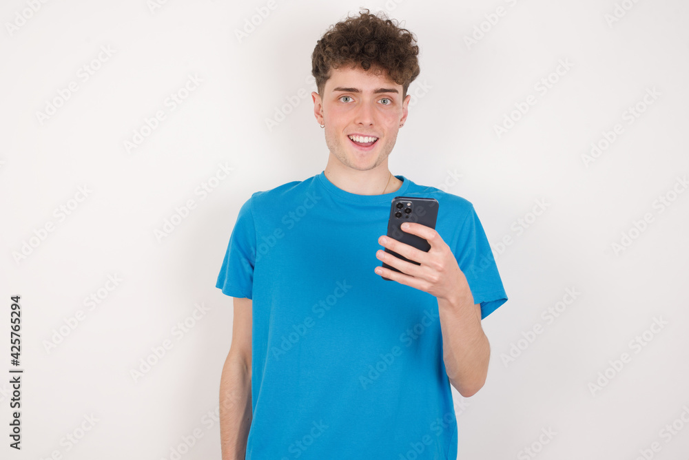 young caucasian handsome man with curly hair wearing blue T-shirt against white studio background holds mobile phone in hands and rejoices positive news, uses modern cellular