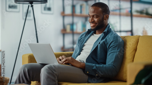 Handsome Black African American Man Working on Laptop Computer while Sitting on a Sofa in Cozy Living Room. Freelancer Working From Home. Browsing Internet, Using Social Networks, Having Fun in Flat.