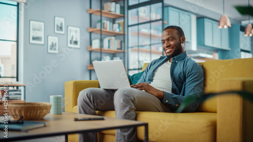 Happy Black African American Man Working on Laptop Computer while Sitting on a Sofa in Cozy Living Room. Freelancer Working From Home. Browsing Internet, Using Social Networks, Having Fun in Flat.
