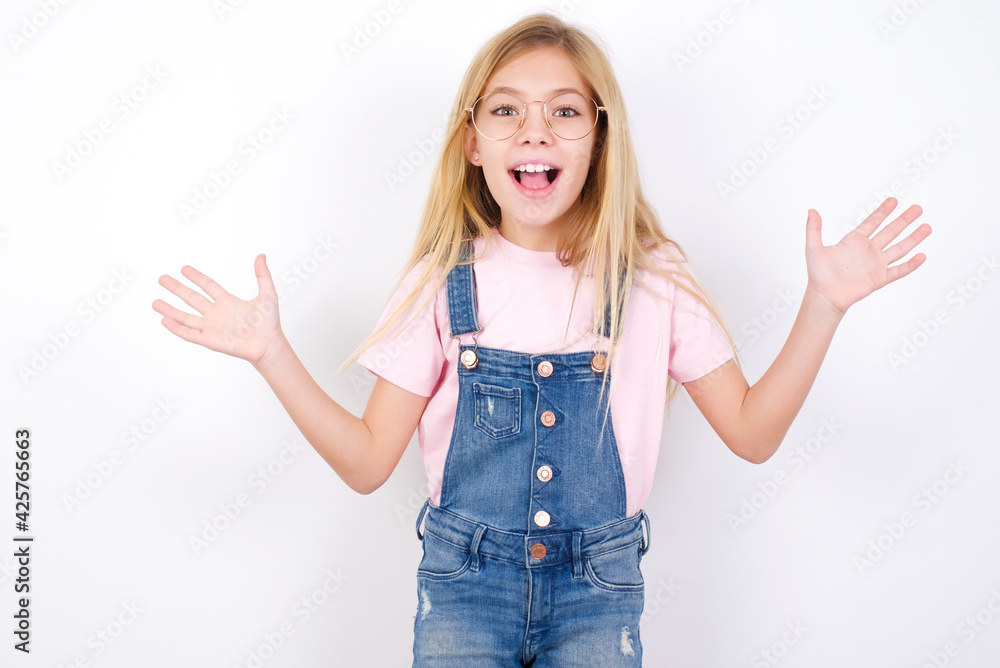 Optimistic beautiful caucasian little girl wearing jeans overall over white background raises palms from joy, happy to receive awesome present from someone, shouts loudly, Excited model screaming. Optimistic beautiful caucasian little girl wearing jeans overall over white background raises palms from joy, happy to receive awesome present from someone, shouts loudly, Excited model screaming.