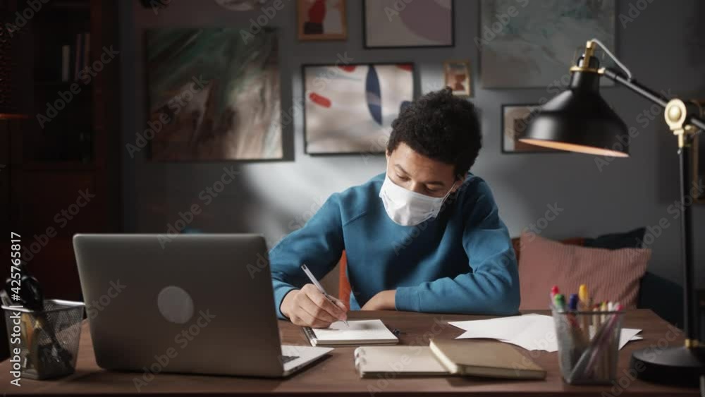 Black African boy student in medical mask doing homework on video conference call, writing notes in copybook. Remote home video lesson. Home isolation, coronavirus quarantine. Black African boy student in medical mask doing homework on video conference call, writing notes in copybook. Remote home video lesson. Home isolation, coronavirus quarantine.