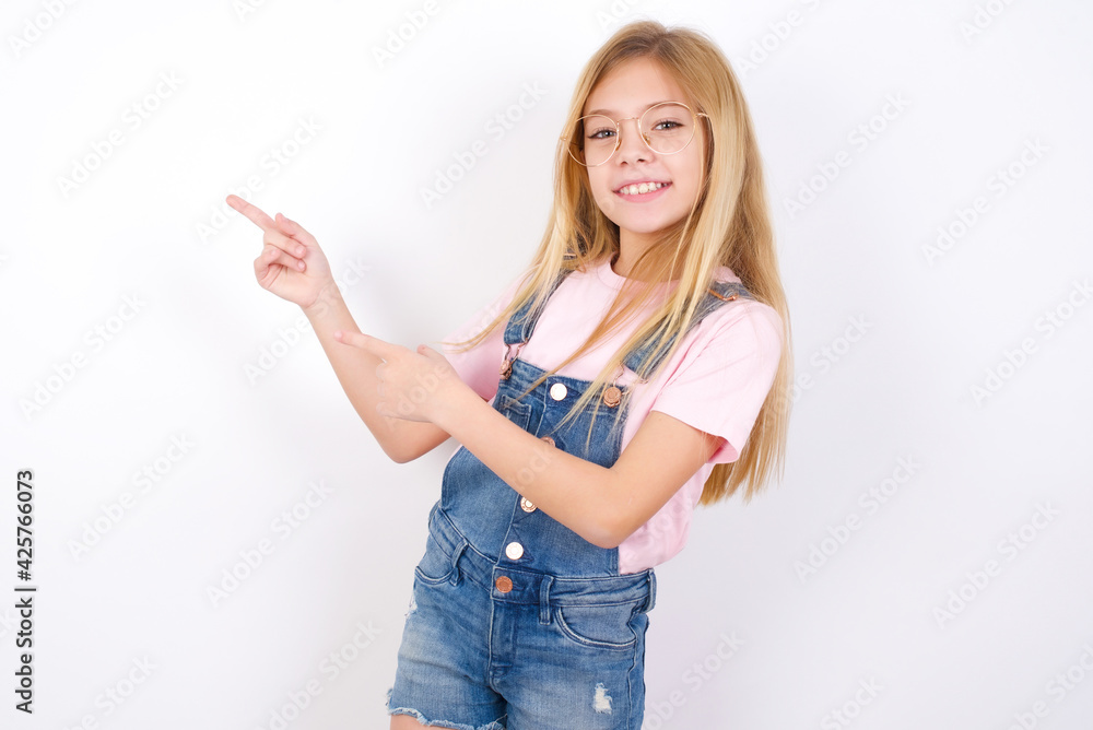 Obraz premium beautiful caucasian little girl wearing jeans overall over white background indicating finger empty space showing best low prices, looking at the camera