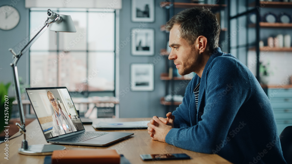 Handsome Caucasian Man Having a Video Call with Female Doctor on Laptop Computer while Sitting Behind Desk in Living Room. Freelancer Working From Home and Talking a Medic Over the Internet. Handsome Caucasian Man Having a Video Call with Female Doctor on Laptop Computer while Sitting Behind Desk in Living Room. Freelancer Working From Home and Talking a Medic Over the Internet.