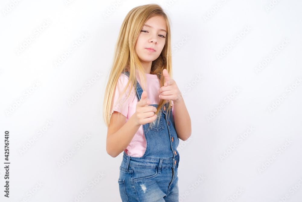 Joyful beautiful caucasian little girl wearing denim jeans overall over white background wink and points index fingers at camera, chooses someone, makes finger gun pistol. Joyful beautiful caucasian little girl wearing denim jeans overall over white background wink and points index fingers at camera, chooses someone, makes finger gun pistol.