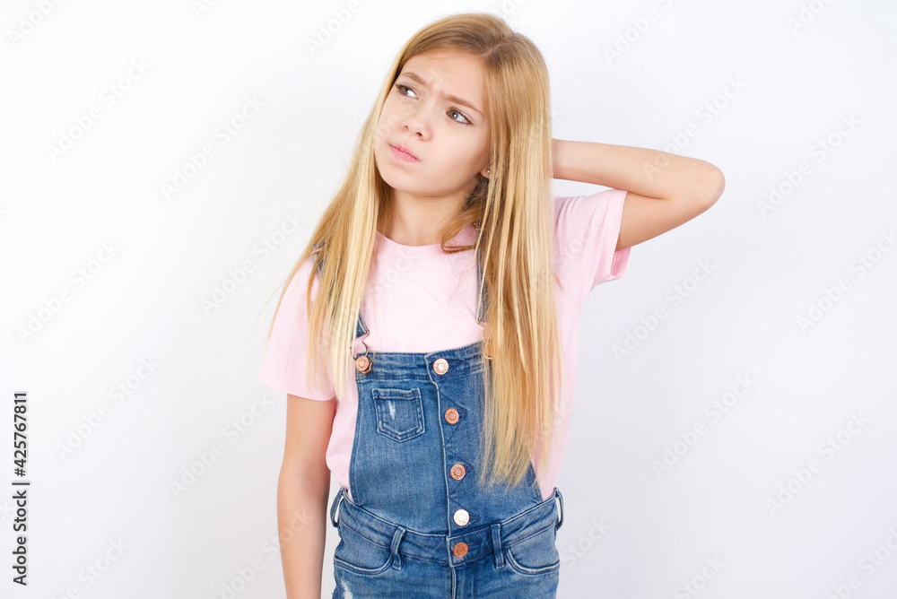 beautiful caucasian little girl wearing denim jeans overall over white background confuse and wonder about question. Uncertain with doubt, thinking with hand on head. Pensive concept. beautiful caucasian little girl wearing denim jeans overall over white background confuse and wonder about question. Uncertain with doubt, thinking with hand on head. Pensive concept.