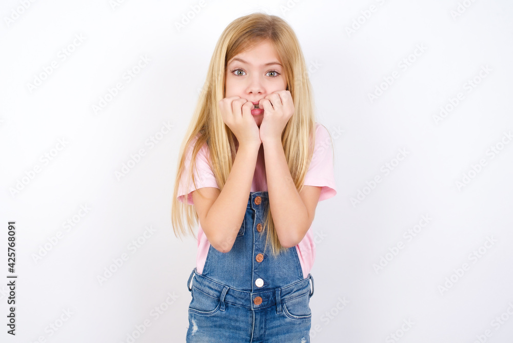 Fearful beautiful caucasian little girl wearing denim jeans overall over white background keeps hands near mouth, feels frightened and scared, has a phobia, Shock and frighted concept. Fearful beautiful caucasian little girl wearing denim jeans overall over white background keeps hands near mouth, feels frightened and scared, has a phobia, Shock and frighted concept.