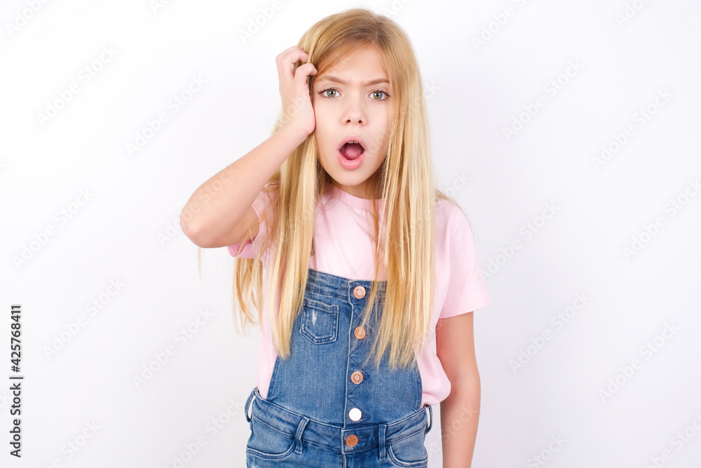 Embarrassed beautiful caucasian little girl wearing denim jeans overall over white background with shocked expression, expresses great amazement, Puzzled model poses indoor Embarrassed beautiful caucasian little girl wearing denim jeans overall over white background with shocked expression, expresses great amazement, Puzzled model poses indoor