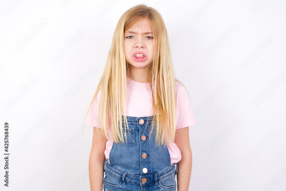 beautiful caucasian little girl wearing denim jeans overall over white background keeps teeth clenched, frowns face in dissatisfaction, irritated because of much duties. beautiful caucasian little girl wearing denim jeans overall over white background keeps teeth clenched, frowns face in dissatisfaction, irritated because of much duties.