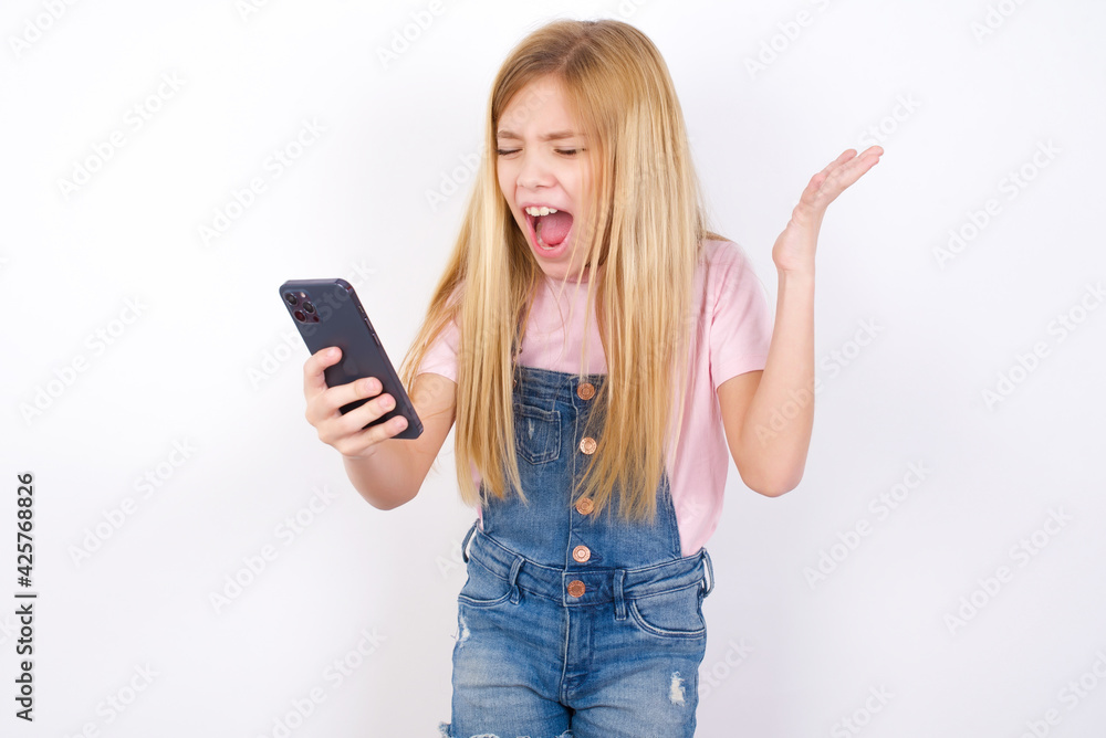 Angry beautiful caucasian little girl wearing denim jeans overall over white background screaming on the phone, having an argument with an employee. Troubles at work. Angry beautiful caucasian little girl wearing denim jeans overall over white background screaming on the phone, having an argument with an employee. Troubles at work.