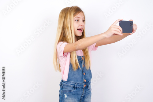 beautiful caucasian little girl wearing denim jeans overall over white background taking a selfie to post it on social media or having a video call with friends.
