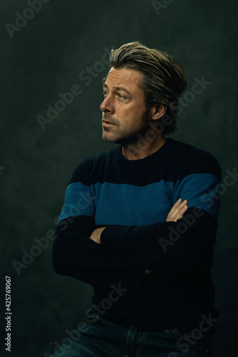 Blond pensive man with arms crossed and stubble beard in blue striped wool sweater in front of a dark wall. Looking along the camera.