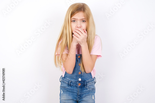 beautiful caucasian little girl wearing denim jeans overall over white background holding oneself  feels very cold outside  hopes that will not get cold